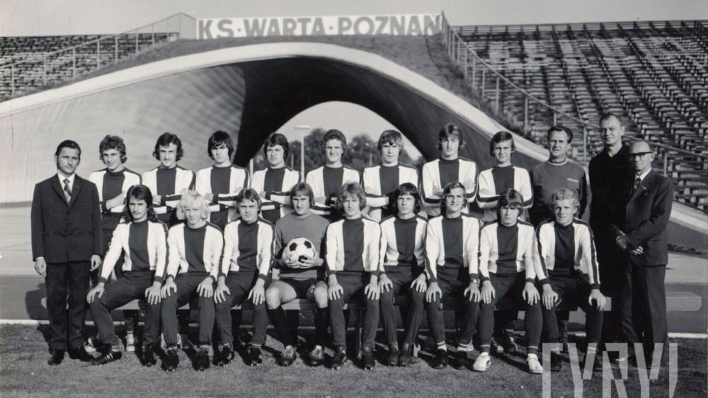 Czarno-białe zdjęcie grupowe zawodników piłkarskich. W tle trybuny Klubu Sportowego Warta Poznań.