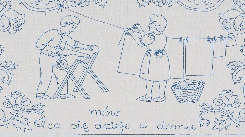 Na makatce mąż piłuje kawałek drewna, a żona obok wiesza pranie na sznurku. Oboje uśmiechnięci.