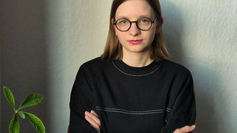 Młoda kobieta ma jasne włosy do ramion i okulary w okrągłych oprawkach, ubrana w czarną bluzę.