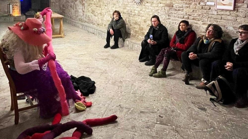 Kobieta w kapeluszu bawi się pluszakami. Patrzą na nią ludzie siedzący naprzeciwko.