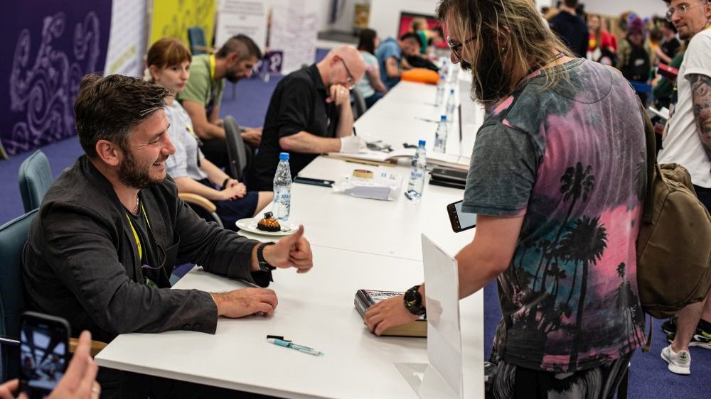 Rząd stolików, za którymi siedzą autorzy rozdający autografy fanom.