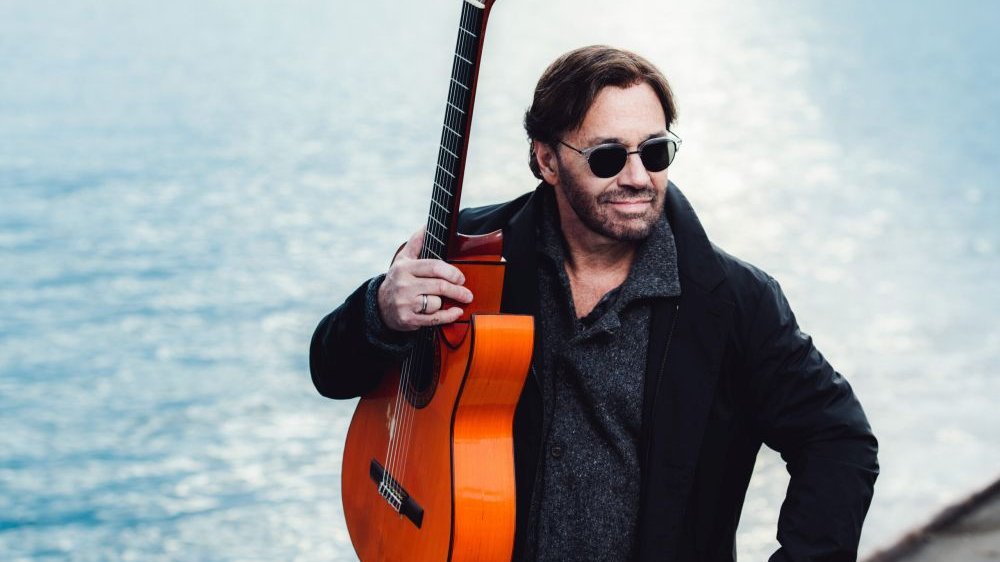 Mężczyzna w okularach przeciwsłonecznych trzyma gitarę stojąc na tle jeziora.