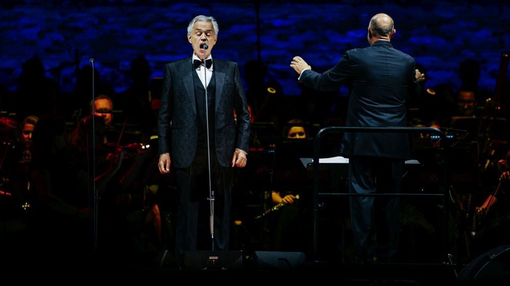 Tenor śpiewa do mikrofonu, za nim dyrygent i orkiestra.