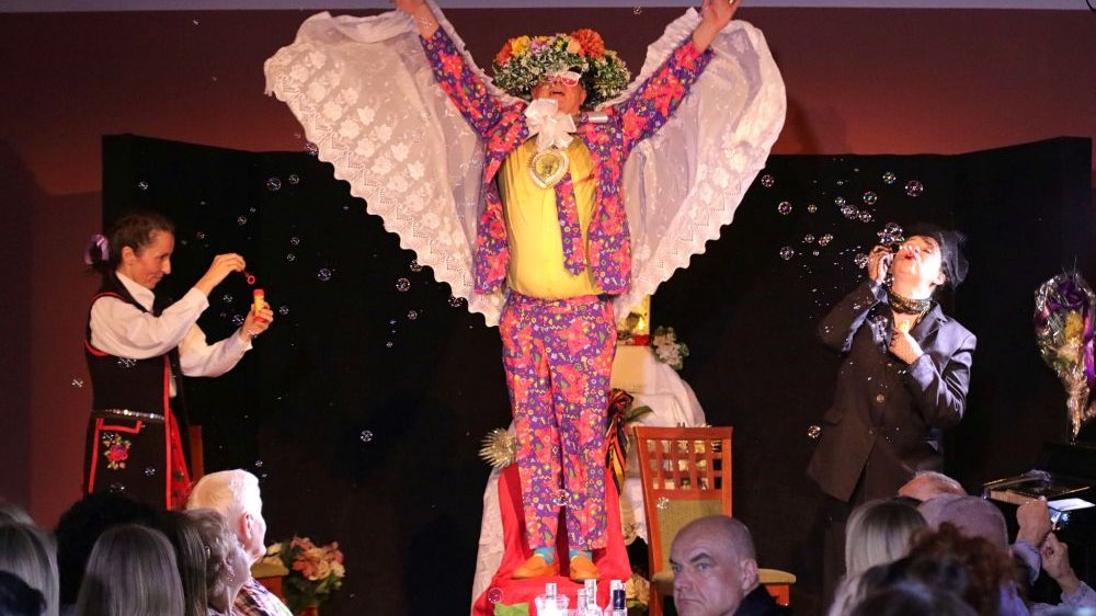 Mała scena, na środku, na krześle stoi mężczyzna w kolorowym garniturze i wianku na głowie, trzyma w ramionach biały oburs lub welon, obok inni dmuchają bańki.