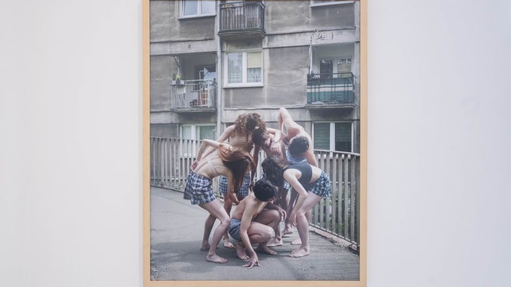 Fotografia na ścianie galerii przedstawiająca grupę ludzi na tle szarego blokowiska.