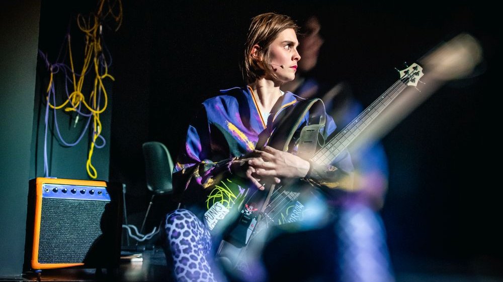 Kolorowo ubrana dziewczyna z gitarą na kolanach patrzy w bok.