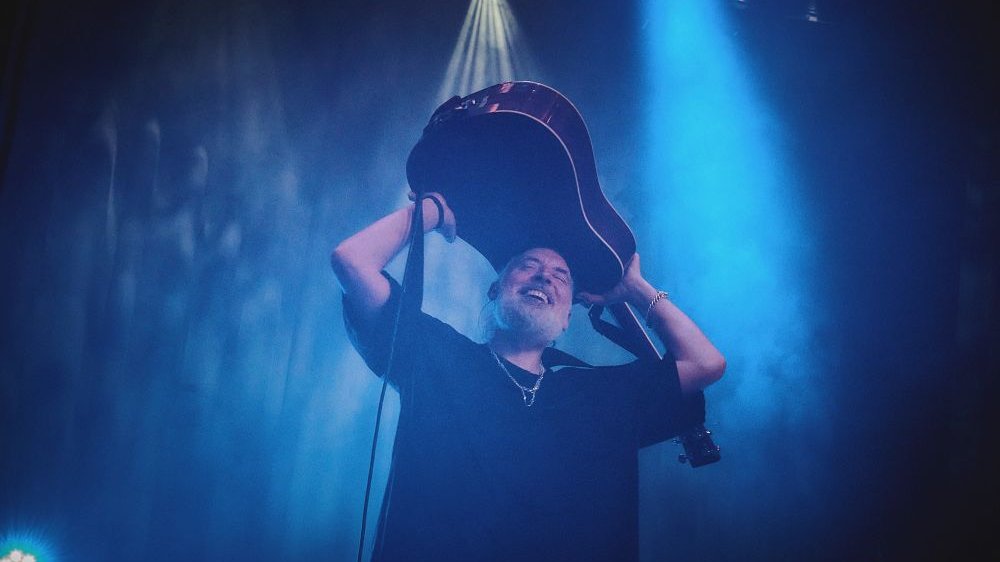 Roześmiany mężczyzna trzyma gitarę nad głową.