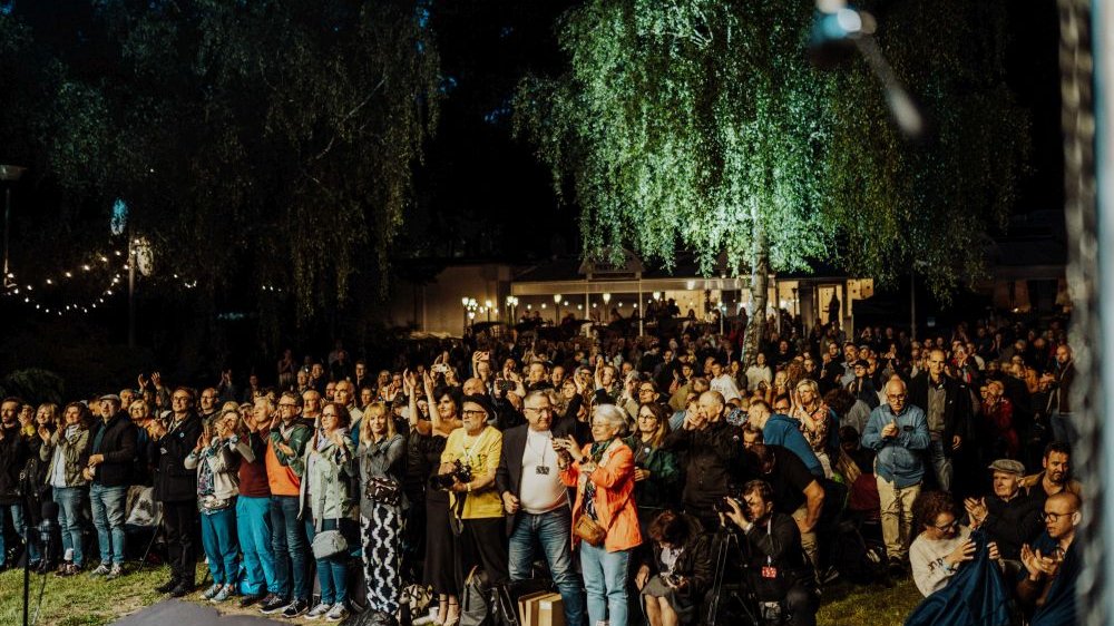 Festiwalowa publiczność stoi, klaszcze. Jest ciemno.