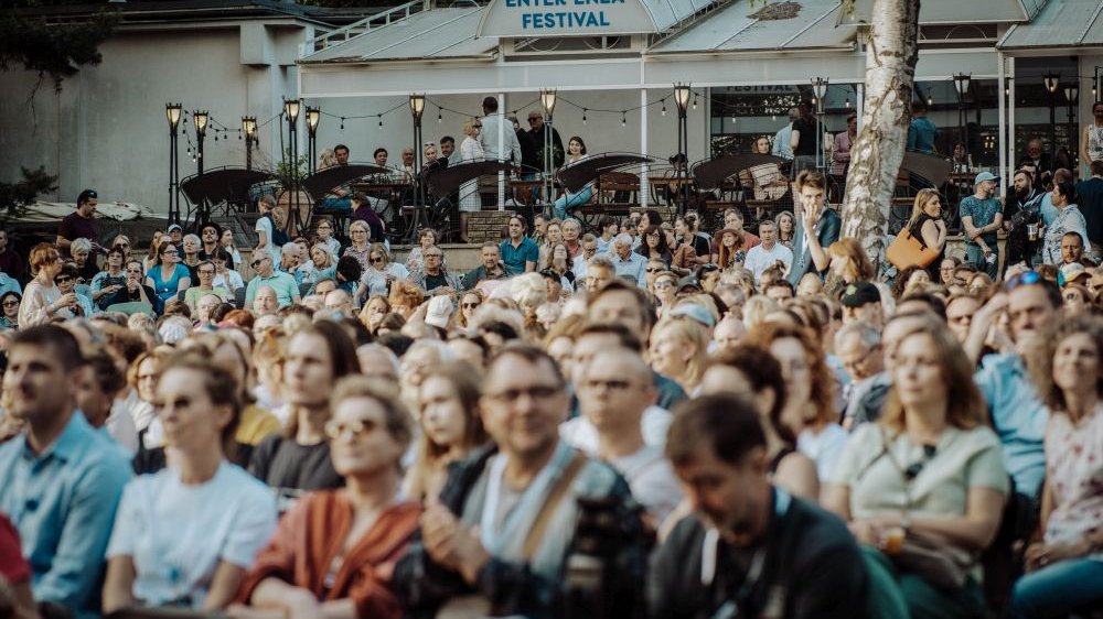 Publiczność festiwalu, w tle bialy pawilon z jego niebieską nazwą.