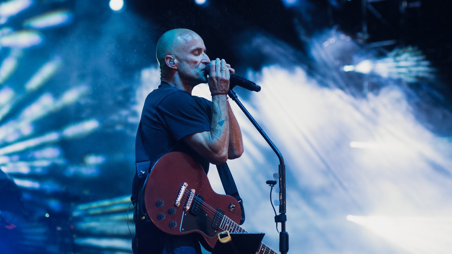 Łysy mężczyna przy mikrofonie, z gitarą na scenie.