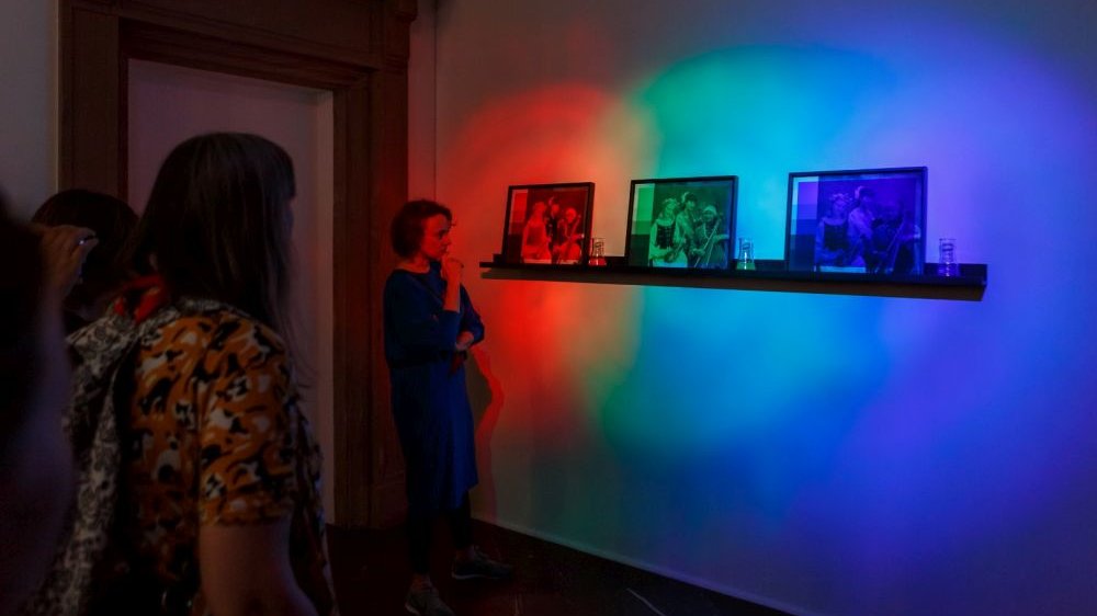 Na ścianie podświetlonej ostrymi światłami - czerwonym, zielonym, niebieskim, jest półka z trzema fotografiami, pozornie do siebie podobnymi. Patrzą na nie trzy zwiedzające.