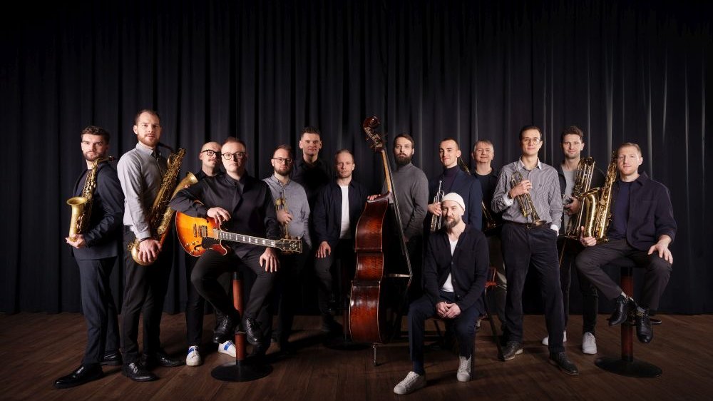 Mężczyźni są młodzi lub w średnim wieku, stoją lub siedzą na scenie, każdy ze swoim instrumentem, za nimi czarna kurtyna.