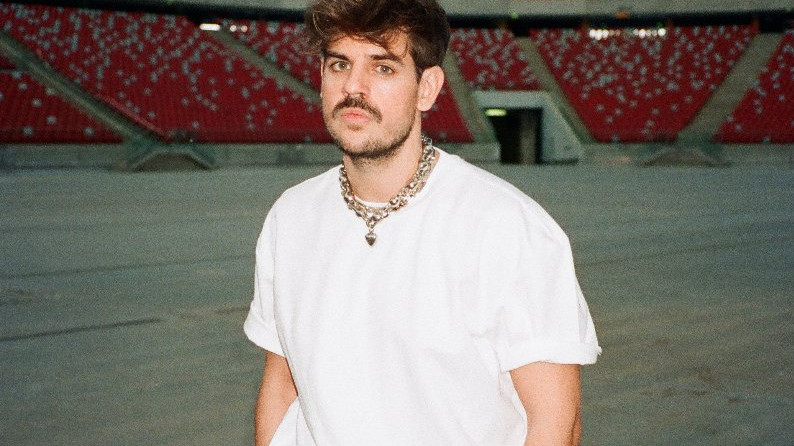 Młody mężczyzna z wąsek i rękoma w kieszeniach spodni pozuje na stadionie. Ma poważną minę.