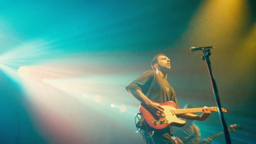 Scena mocno oświetlona błękitnym i złotym światłem, przy mikrofonie chłopak grający na gitarze elektrycznej.