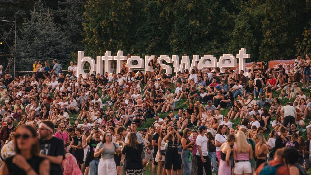 Tłum ludzi siedzi na trawie na tle stojącego napisu z nazwą festiwalu.