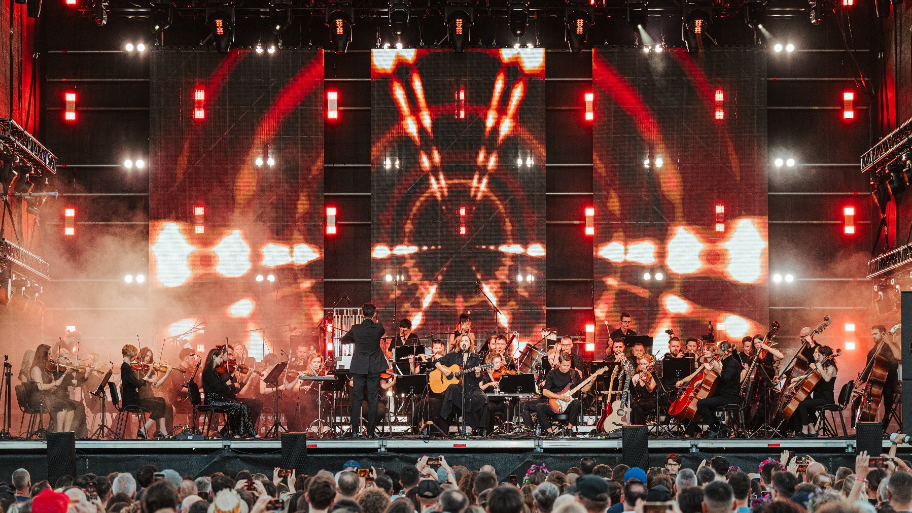 Orkiestra, za nią wizualizacja na całejk scenie. W centrum główna artystka z gitarą.