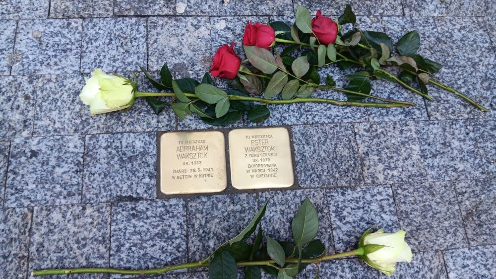 Złote kamienie z imionami i nazwiskami wmurowane w chodnik. Obok nich leżą białe i czerwone róże.