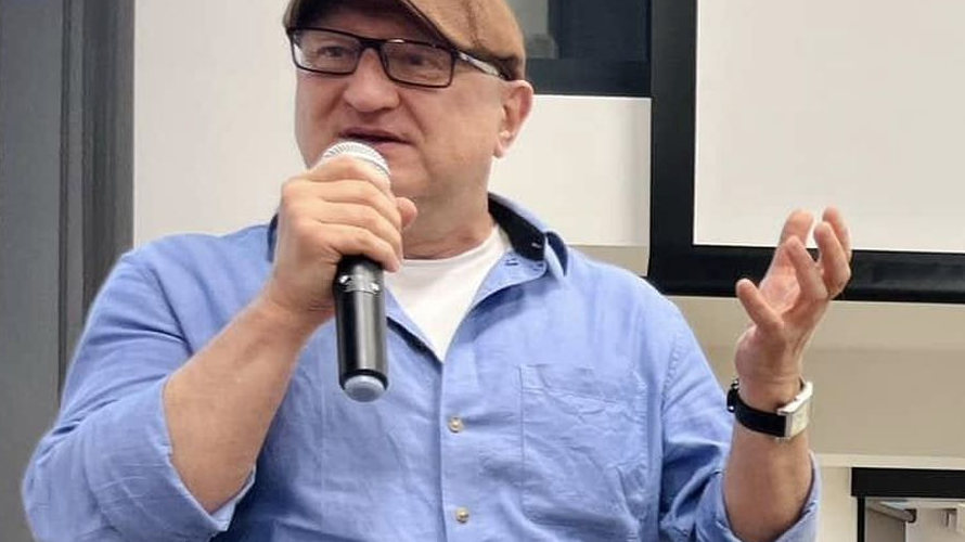 Mężczyzna w kaszkiecie i błękitnej koszuli opowiada coś do mikrofonu.