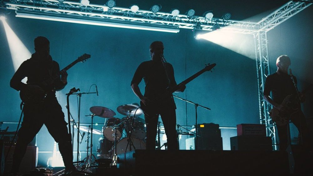 Trzy postaci w cieniu sceny grają rockowy koncert.