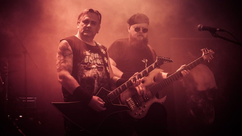 Dwaj mężczyźni grają na elektrycznych gitarach na zadymionej scenie.