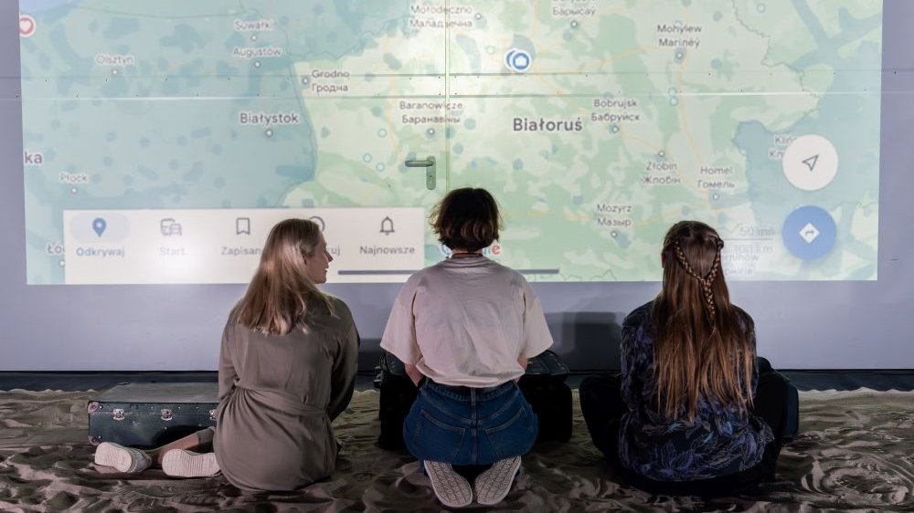 Trzy dziewczyny klęczą na piasku patrząc na duży ekran z mapą Białorusi.