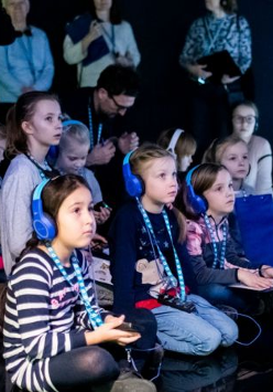 Dzieci siedzące na podłodze w słuchawkach. Za nimi rodzice/opiekunowie. Wszyscy znajdują sie w sali multimedialnej. Na podłodze wywietlają się animacje.