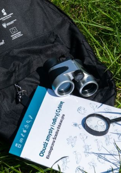 czarny pleacak leżący na trawie. Na nim lornetka, lupa i zeszyt.