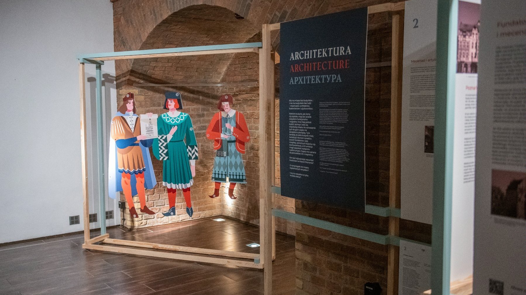 Część ekspozycji poświęcona architekturze. Drewniane sylwetki trzech ludzi w dawnych strojach podwieszone w ramie, obok tablica z tekstem.