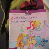 Okładka książki. Dziwczynka z kokardą we włosach przymierza stroje na bal.