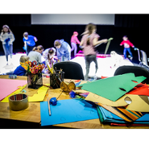 na zdjeciu grupa dzieci podczas zajęć. W tle widać siedzące na podłodze i biegajace dzieci. Na pierwszym planie stół z przyborami do zajęć plastycznych: kredkami, kolorowym papierem, nożyczkami.