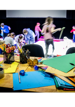 na zdjeciu grupa dzieci podczas zajęć. W tle widać siedzące na podłodze i biegajace dzieci. Na pierwszym planie stół z przyborami do zajęć plastycznych: kredkami, kolorowym papierem, nożyczkami.