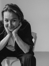 Zdjęcie młodej kobiety, która uśmiechnięta siedzi na krześle, rekoma podpiera głowę.