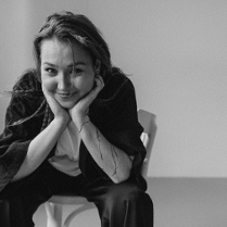 Zdjęcie młodej kobiety, która uśmiechnięta siedzi na krześle, rekoma podpiera głowę.