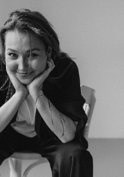 Zdjęcie młodej kobiety, która uśmiechnięta siedzi na krześle, rekoma podpiera głowę.