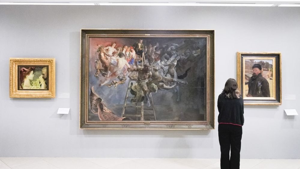 Obrazy Jacka Malczewskiego na ścianie Muzeum, ogląda je kobieta w czerni. Obraz na środku zdecydowanie góruje rozmiarami nad pozostałymi obrazami po swojej prawej i lewej stronie.