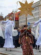 Zdjęcie przedstawia tłum ludzi idących ulicą. W pierwszym rzędzie widać dwie dziewczyny idące na szczudłach w przebraniu aniołów oraz mężczyznę trzymającego w ręku dużą złotą gwiazdę.