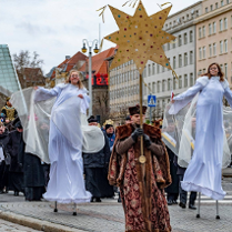 Zdjęcie przedstawia tłum ludzi idących ulicą. W pierwszym rzędzie widać dwie dziewczyny idące na szczudłach w przebraniu aniołów oraz mężczyznę trzymającego w ręku dużą złotą gwiazdę.