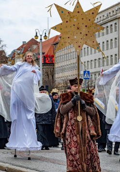Zdjęcie przedstawia tłum ludzi idących ulicą. W pierwszym rzędzie widać dwie dziewczyny idące na szczudłach w przebraniu aniołów oraz mężczyznę trzymającego w ręku dużą złotą gwiazdę.