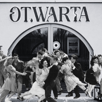 Zdjęcie przedstawia restaurację z parami oddającymi się tańcowi swingowemu. Dziewczyny w rękach swoich partnerów latają i śmieją się.