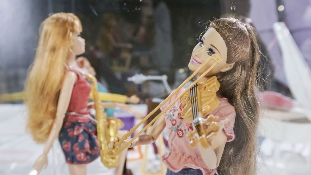 Dwie lalki Barbie na wystawie. Lalka z przodu gra na skrzypcach, lalka z tyłu trzyma w rękach saksofon.