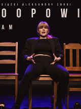 Kobieta w jasnych włosach z grzywką. Ubrana na czarno. Siedzi na jednym z trzech krzeseł, na środku. Z góry świeci na nią różowe światło. Tło jest ciemne.