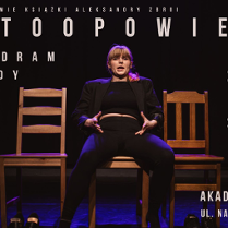 Kobieta w jasnych włosach z grzywką. Ubrana na czarno. Siedzi na jednym z trzech krzeseł, na środku. Z góry świeci na nią różowe światło. Tło jest ciemne.