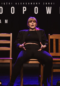 Kobieta w jasnych włosach z grzywką. Ubrana na czarno. Siedzi na jednym z trzech krzeseł, na środku. Z góry świeci na nią różowe światło. Tło jest ciemne.