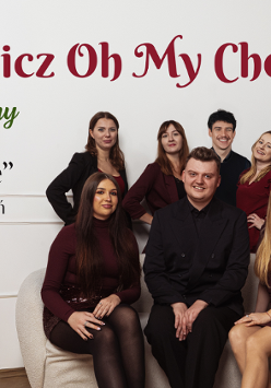 Zespół muzyczny składający się z dwóch mężczyzn i siedmiu kobiet. Mężczyźni ubrani w czarne koszule i spodnie. Kobiety ubrane w bordowe sukienki. Wszyscy się uśmiechają i pozują do zdjęcia na tle przystrojonej choinki.