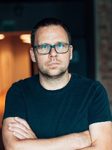 Zdjęcie przedstawia mężczyznę w okularach. Stoi z rękami złożonymi na piersi. Jego spojrzenie jest poważne.