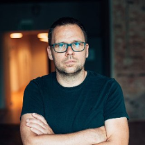 Zdjęcie przedstawia mężczyznę w okularach. Stoi z rękami złożonymi na piersi. Jego spojrzenie jest poważne.