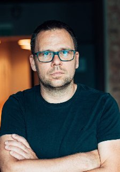 Zdjęcie przedstawia mężczyznę w okularach. Stoi z rękami złożonymi na piersi. Jego spojrzenie jest poważne.