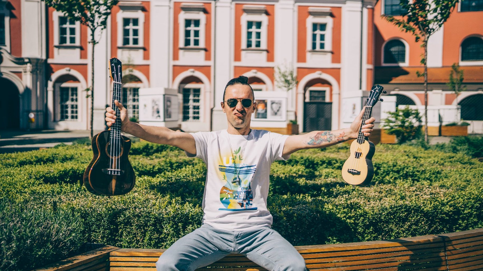 Rozmówca w białej koszulce na dziedzińcu Urzędu Miasta Poznania, trzyma w rękach dwa instrumenty ukulele, przypominające małe gitary