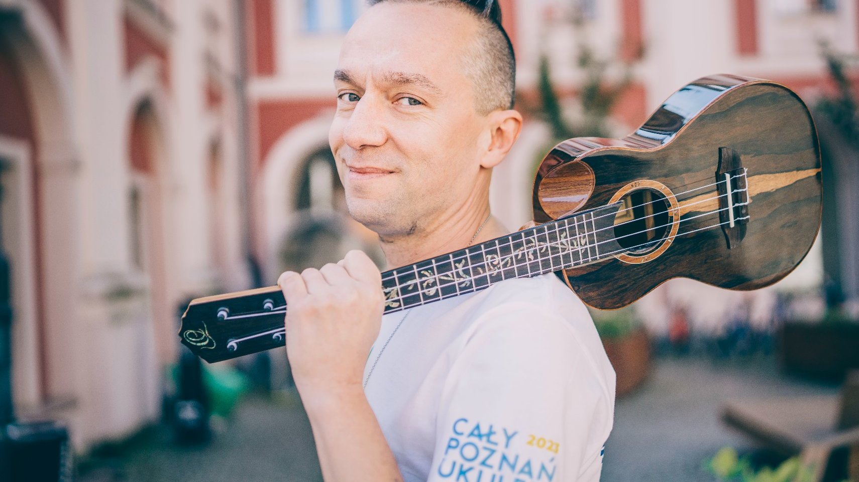 Mężczyzna w białej koszulce na dziedzińcu Urzędu Miasta Poznania, trzyma w rękach, zwrócone w strone obiektywu, ukulele. Instrument przypomina małą gitarę, ma 4 struny