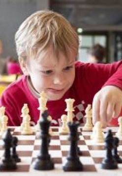 Chłopiec w czerwonej koszulce gra w szachy, za nim bawią się inne dzieci.